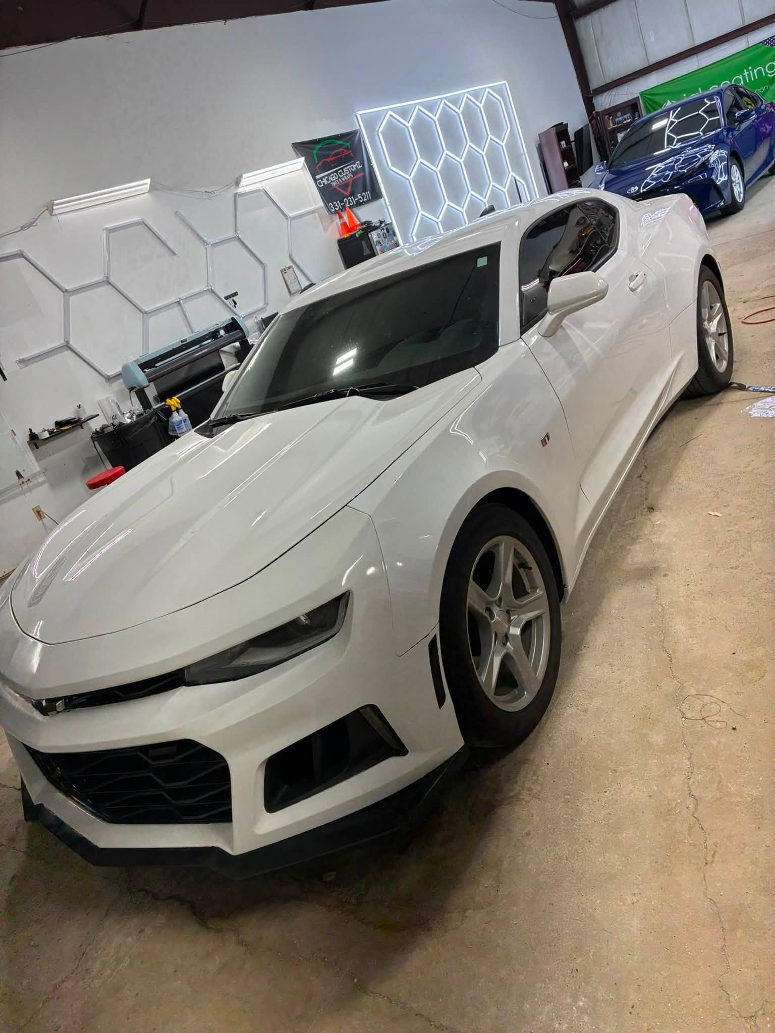 White Chevrolet Camaro with tinted windows in front of the shop's hexagon light panel